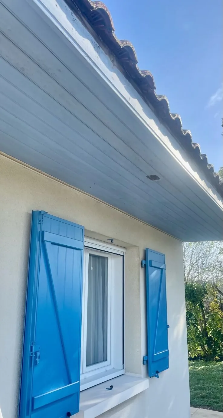 Plafond de débord de toit taché avant peinture, au-dessus d'une fenêtre à volets bleus